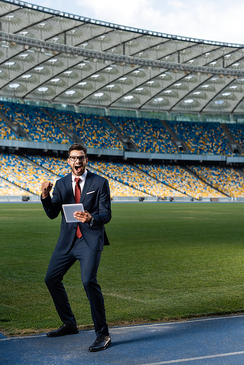 young businessman in suit and glasses using digital tablet and showing yeas gesture at stadium, sports betting concept