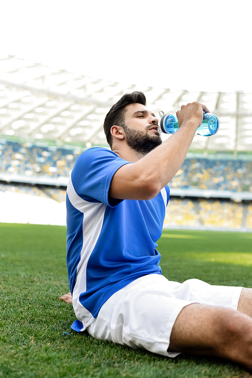 professional soccer player in blue and white uniform sitting on football pitch and drinking water at stadium