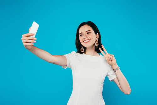 smiling elegant woman in dress taking selfie and showing peace sign isolated on blue