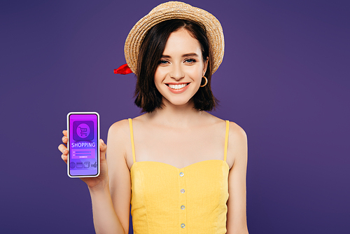 smiling girl in straw hat holding smartphone with shopping app isolated on purple