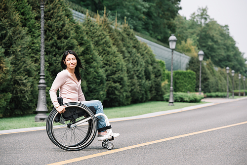 beautiful disabled woman in wheelchair smiling at camera while walking in park