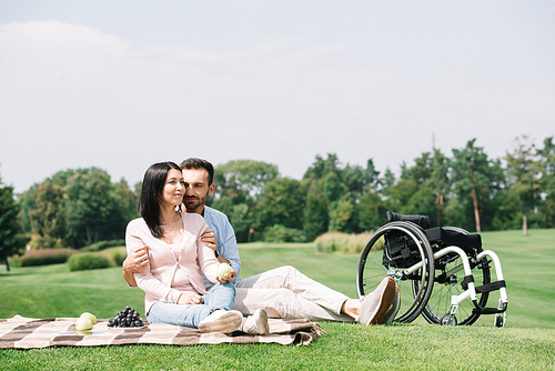 happy handsome man hugging young disabled girlfriend while sitting on blanket in park