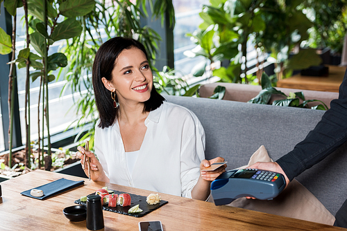 cropped view of waiter holding credit card reader near attractive woman paying with credit card