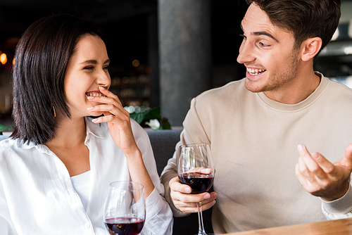 happy man looking at woman laughing while holding glasses with red wine