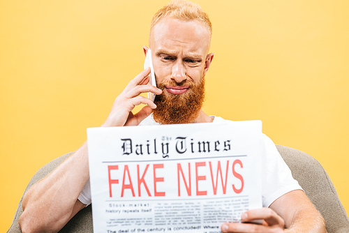 skeptical man reading newspaper with fake news and talking on smartphone while sitting on armchair, isolated on yellow