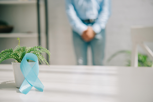 selective focus of blue awareness ribbon near flowerpot and man standing by wall and holding clenched hands near groin area
