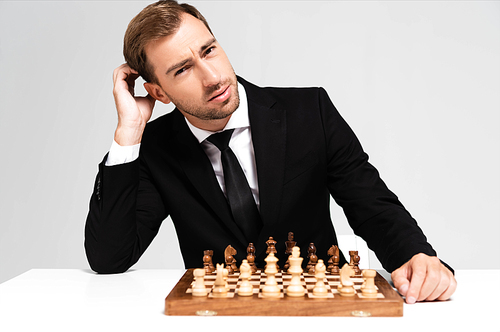 pensive and handsome businessman in suit sitting near chessboard isolated on grey
