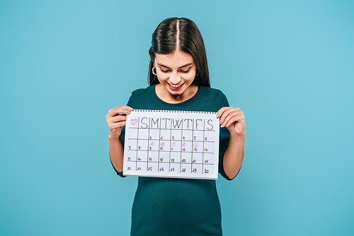 smiling pregnant girl looking at period calendar isolated on blue