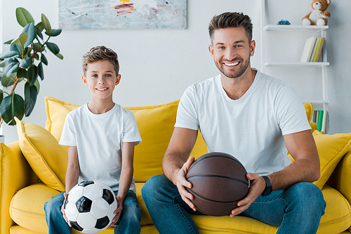 happy man holding basketball near son with football at home