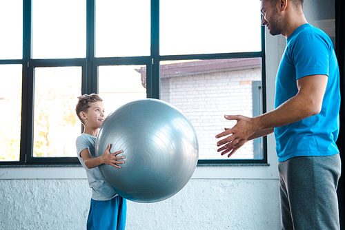 cute kid holding fitness ball and looking at father in gym