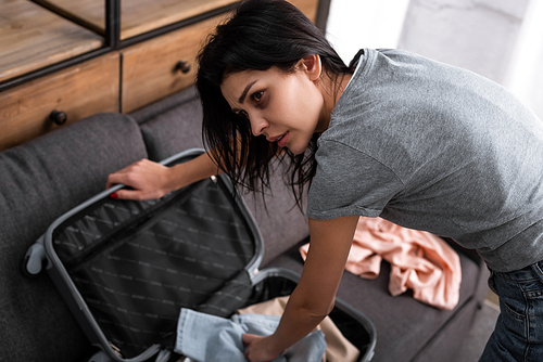 selective focus of sad woman with bruise on face packing suitcase with clothing, domestic violence concept