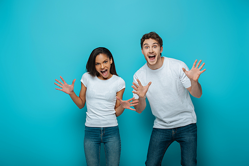 cheerful interracial couple showing frightening gestures at camera on blue background