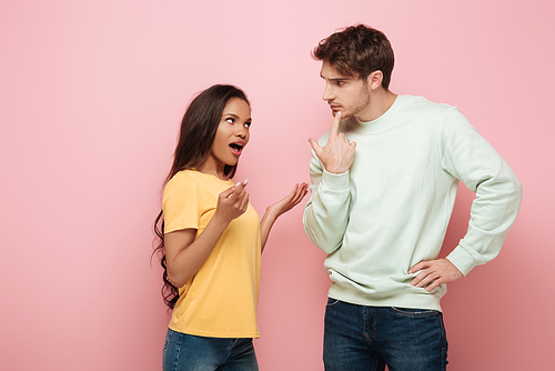 serious guy touching chin while looking at african american girlfriend quarreling on pink background