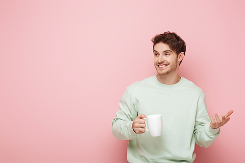 cheerful young man holding cup while standing with open arm and looking away on pink background