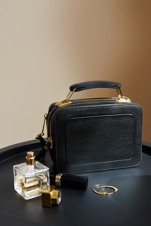 leather handbag near golden earrings, perfume and lipstick on black table on beige background