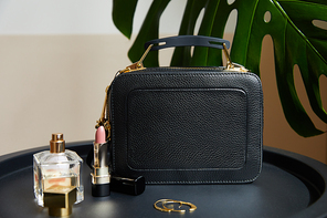 leather handbag near golden earrings, perfume and lipstick on black table near tropical leaf