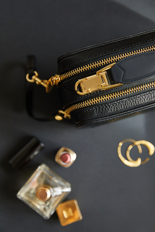 top view of leather handbag near golden earrings, perfume and lipstick on black table