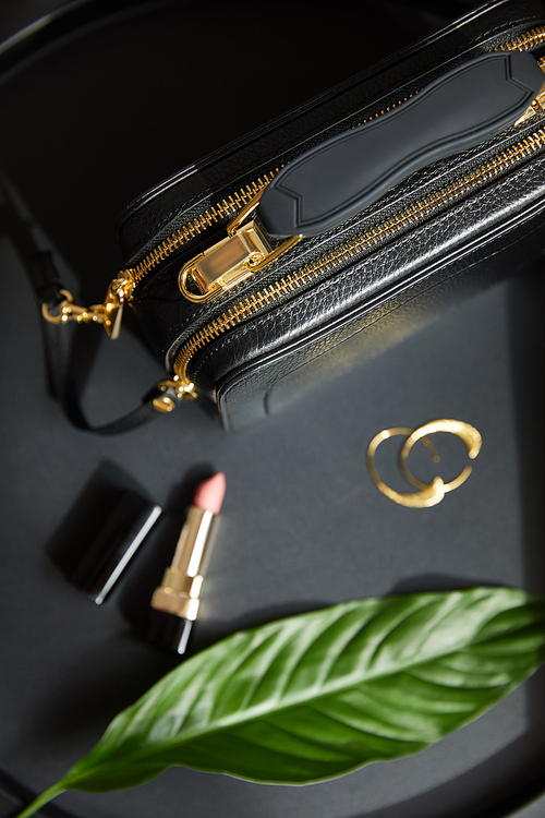 top view of leather handbag near golden earrings and lipstick on black table with green leaf