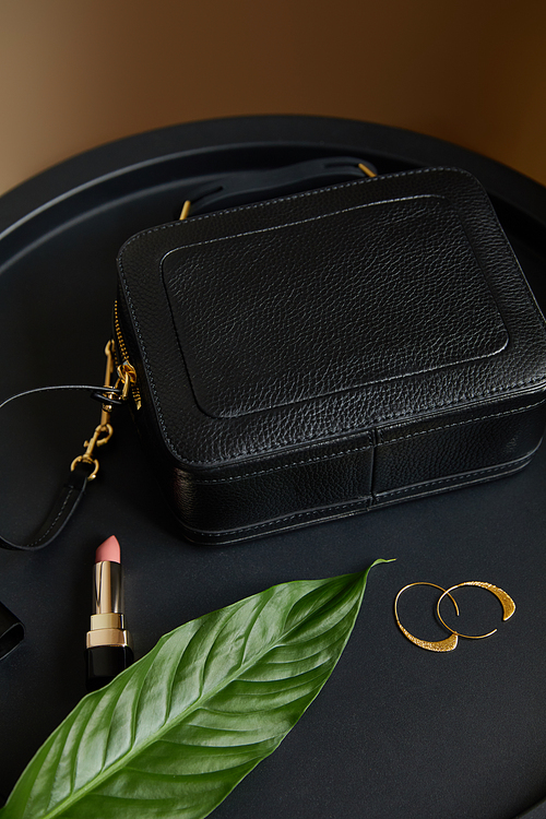 leather handbag near golden earrings and pink lipstick on black table with tropical leaf