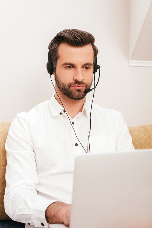 Selective focus of handsome teleworker using laptop and headset at home