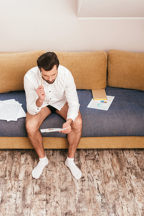 Pensive freelancer in shirt and panties looking at documents near book on sofa in living room