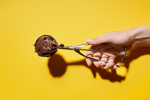 partial view of woman holding fresh chocolate ice cream ball in scoop on yellow background