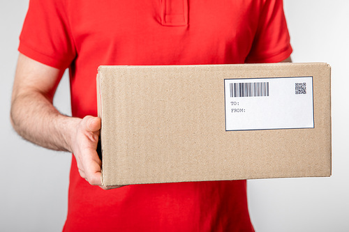 Cropped view of courier carrying cardboard box with barcode and qr code on card isolated on grey