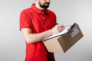 Cropped view of delivery man writing on clipboard and holding cardboard box isolated on grey