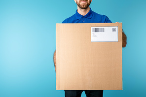 Cropped view of courier holding cardboard box with qr and barcode on card isolated on blue