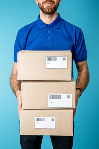 Cropped view of courier holding cardboard boxes with qr codes and barcodes on cards isolated on blue