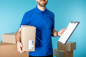 Cropped view of delivery man holding cardboard box and clipboard with copy space near boxes isolated on blue