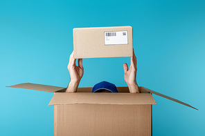 Cropped view of courier in cap holding cardboard package from box isolated on blue