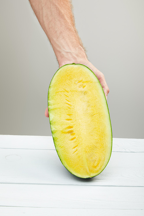cropped view of man holding ripe yellow watermelon half on wooden table isolated on grey