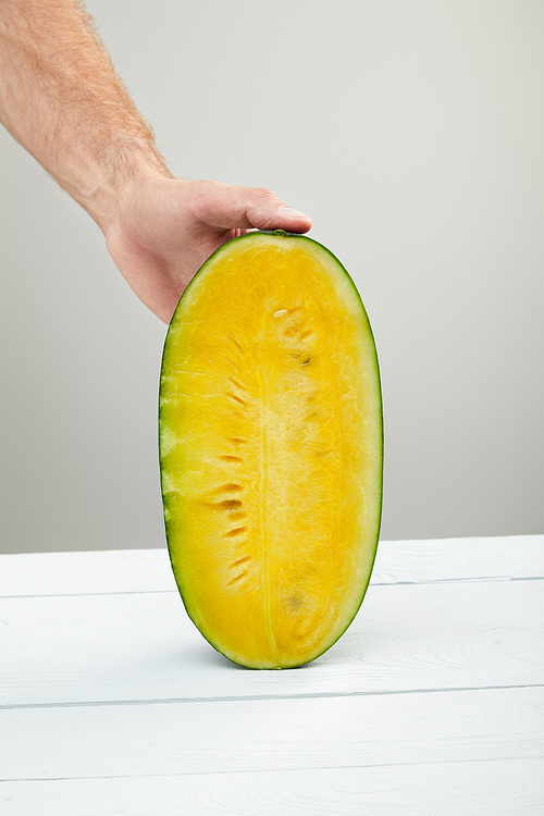 partial view of man holding ripe yellow watermelon half on wooden table isolated on grey