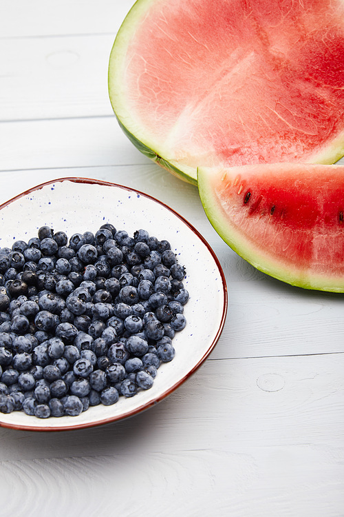 cut ripe red watermelon with seeds near plate with blueberries on wooden white table isolated on grey
