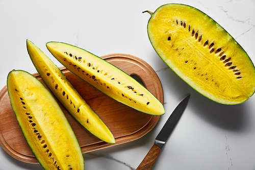 top view of cut delicious exotic yellow watermelon with seeds on wooden chopping board with knife on marble surface