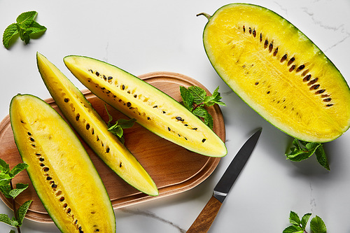 top view of cut delicious exotic yellow watermelon with seeds on wooden chopping board with knife and mint on marble surface