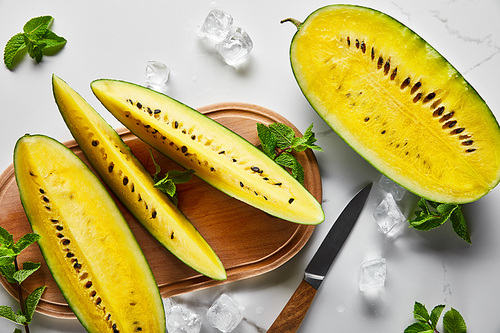 top view of cut delicious exotic yellow watermelon with seeds on wooden chopping board with knife, mint and ice cubes on marble surface