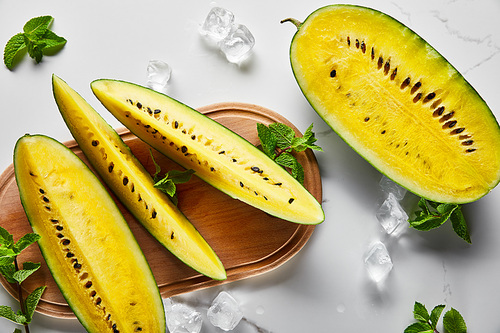 top view of cut delicious exotic yellow watermelon with seeds on marble surface with mint, ice and wooden chopping board