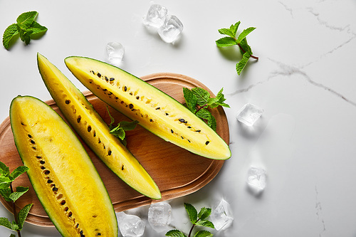 top view of cut exotic yellow watermelon with seeds on marble surface with mint, ice and wooden chopping board