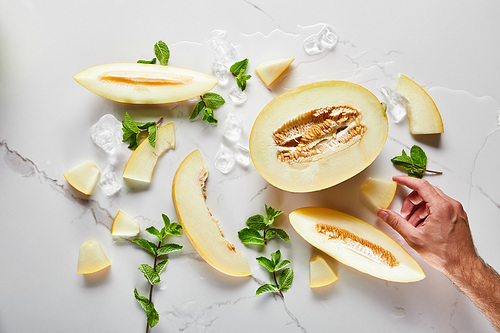 top view of cut delicious melon on marble surface with mint and ice near man hand