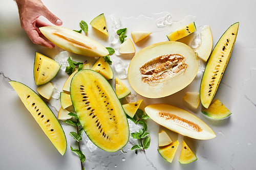 cropped view of man holding cut delicious  near exotic yellow watermelon on marble surface with ice and mint