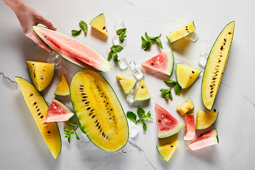 cropped view of hand near delicious exotic yellow and red watermelons with ice and mint on marble surface