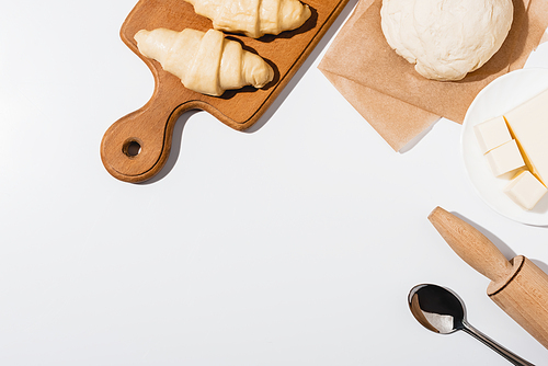 top view of fresh croissants on wooden cutting board near raw dough, rolling pin, butter and spoon on white background
