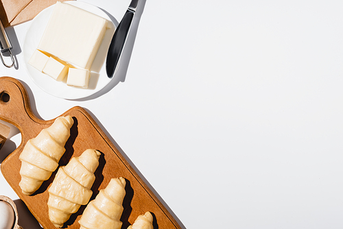 top view of fresh raw croissants on wooden cutting board near butter on plate with knife on white background