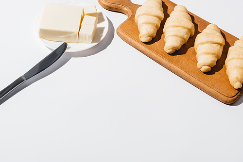fresh raw croissants on wooden cutting board near butter on plate with knife on white background