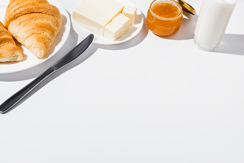tasty fresh baked croissants on plate near glass of milk, butter with knife and jam on white background