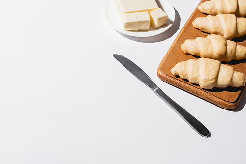 fresh raw croissants on wooden cutting board near butter on plate with knife on white background
