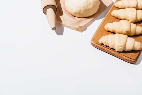 fresh croissants on wooden cutting board near raw dough, rolling pin on white background