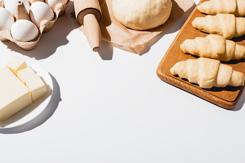 fresh croissants on wooden cutting board near raw dough, rolling pin, butter and eggs on white background
